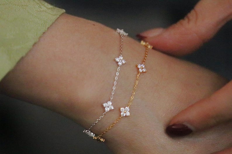 May include: Three delicate bracelets with floral designs. One is gold, one is silver, and the third is rose gold. Each bracelet features small, sparkling flower-shaped accents. The bracelets are worn on a wrist.