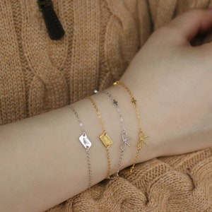 Puede incluir: Un conjunto de cuatro delicados brazaletes de oro y plata con diferentes dijes, incluyendo una estrella, un lazo y una etiqueta rectangular. Los brazaletes están superpuestos en la muñeca de una persona.