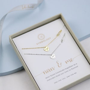 May include: A white box with a gold and silver necklace set inside. The box has a white background with the text "SYMBOLOGY" and "nan & me" printed on it. The text reads "wear these bracelets together as a reminder that our love is forever. ONE FOR YOU, ONE FOR ME."