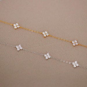May include: Two delicate necklaces, one gold and one silver, each adorned with multiple four-petal flower charms. The charms are encrusted with small, sparkling crystals. The necklaces are displayed on a neutral background.