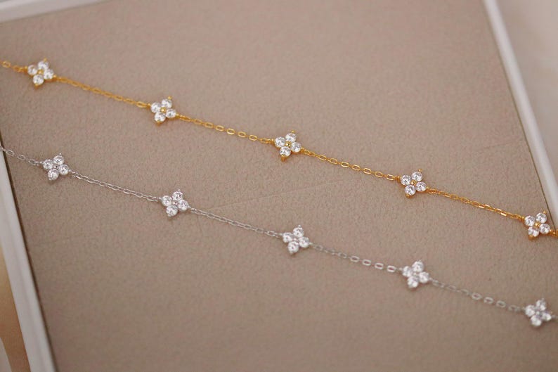 May include: Two delicate necklaces, one gold and one silver, each adorned with multiple four-petal flower charms. The charms are encrusted with small, sparkling crystals. The necklaces are displayed on a neutral background.