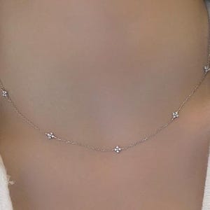 May include: Delicate silver necklace featuring small, sparkling floral-shaped pendants evenly spaced along the chain. The necklace is worn against a white top, highlighting the jewelry's subtle elegance. A refined accessory for everyday wear.