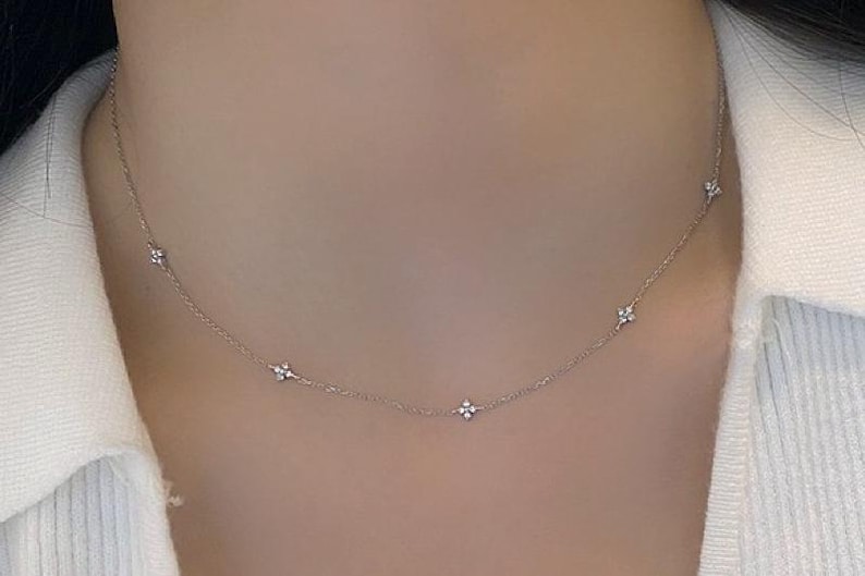 May include: Delicate silver necklace featuring small, sparkling floral-shaped pendants evenly spaced along the chain. The necklace is worn against a white top, highlighting the jewelry's subtle elegance. A refined accessory for everyday wear.
