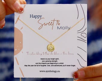 May include: A gold-colored necklace with a round pendant featuring a starburst design, presented on a card. The card reads "Happy Sweet 16 Molly" with additional text about dreams and adventures. The necklace is held by a person.