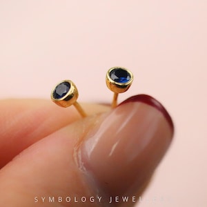 May include: Close-up of two gold stud earrings with round, deep blue gemstones. The earrings are held between fingers against a soft pink background. The earrings have a simple, elegant design.