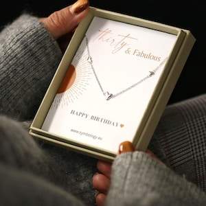 May include: A silver necklace with heart-shaped pendants is displayed in a gift box. The box has the text "thirty & fabulous" and "Happy Birthday". The necklace is on a card with a sunburst design.