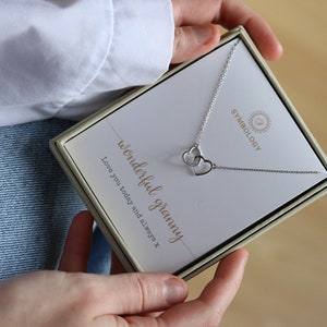 Può includere: Una scatola regalo bianca con un ciondolo d'argento a forma di due cuori intrecciati. La scatola contiene un biglietto bianco con la scritta "wonderful granny" e "Love you today and always x".