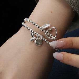 May include: Two silver beaded bracelets with charms. One bracelet features a heart-shaped charm, and the other has a charm with a letter and a butterfly. The bracelets are worn on a wrist.
