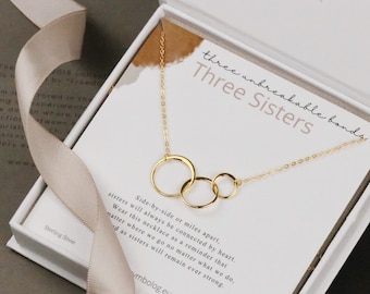 May include: A gold necklace with three interlocking circles, presented in a white box with the text "three unbreakable bonds, Three Sisters" on the lid. The necklace is accompanied by a card with a message about sisters being connected by heart, no matter the distance.