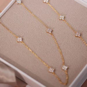 Puede incluir: Collares dorados con cadenas delicadas y detalles florales de piedras preciosas transparentes. Los collares se exhiben en una caja blanca con interior beige, y se ve una cinta de satén.