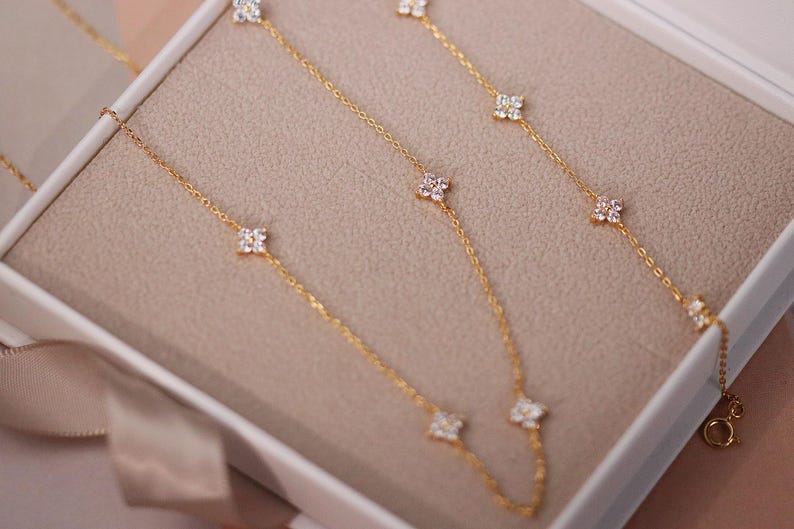 May include: Gold-toned necklaces with delicate chains and floral-shaped, clear gemstone accents. The necklaces are displayed in a white box with a beige interior, and a satin ribbon is visible.