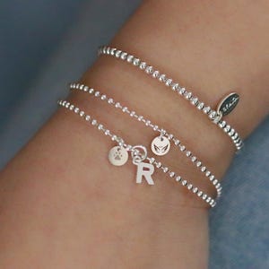 Può includere: Tre braccialetti di perline d'argento sono avvolti attorno a un polso. Due dei braccialetti hanno piccoli ciondoli con un'impronta di zampa, la lettera "R" e un disegno ad ali. Il terzo braccialetto ha una piccola etichetta con la parola "Stella".