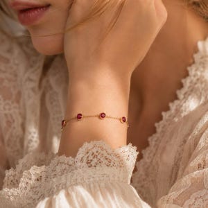 May include: A delicate gold chain bracelet featuring four evenly spaced, round, bezel-set red gemstones. The bracelet is worn on a wrist, showcasing the thin metallic links and the vibrant, polished finish of the circular red stones set in gold.
