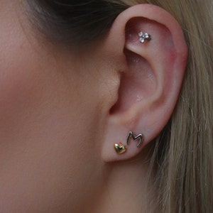 Peut inclure: Gros plan sur une oreille ornée de trois boucles d'oreilles. Une petite boucle d'oreille en forme de cœur doré est dans le lobe inférieur. Une boucle d'oreille en argent en forme de lettre "M" est à côté du cœur. Une petite boucle d'oreille fleur en argent est dans le haut de l'oreille.