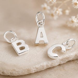 Op de afbeelding: Drie individuele zilverkleurige metalen bedels met de hoofdletters A, B en C. Elke bedel is voorzien van een kleine ronde ring aan de bovenkant voor bevestiging en heeft een gladde, gepolijste afwerking in een strak lettertype.