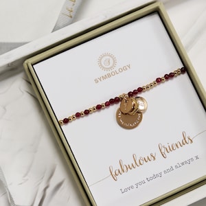 Op de afbeelding: Een gouden armband met rode kralen en drie gouden bedels. De armband zit in een witte doos met een gouden en witte kaart waarop "Symbology" en "fabulous friends Love you today and always x" staat.