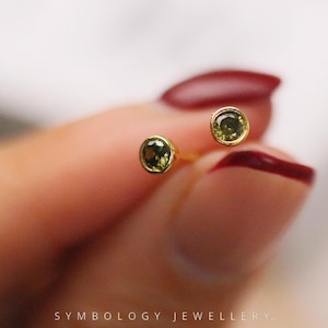 Könnte beinhalten: Ein Paar kleine, runde Ohrstecker mit Goldfassung und einem grünen Edelstein. Die Ohrringe werden vor einem verschwommenen Hintergrund gezeigt, mit dem Text "SYMBOLOLOGY JEWELLERY" unten.