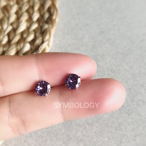 Puede incluir: Un par de pequeños pendientes de botón redondos de color morado. Los pendientes están engastados en plata.