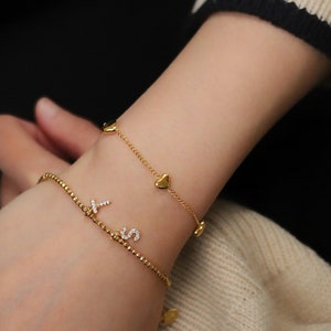 Könnte beinhalten: Zwei goldene Armbänder am Handgelenk einer Person. Ein Armband hat einen Herz-Anhänger und das andere einen Buchstaben-Anhänger.