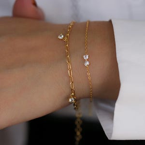 May include: Two gold bracelets on a wrist. One bracelet has a chain link design with three clear, round charms. The other bracelet is a delicate chain with two bow-shaped charms with clear stones.
