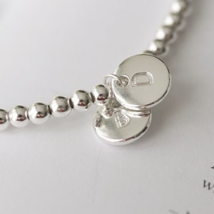 Op de afbeelding: Een zilveren armband met kralen en twee zilveren bedels, elk met een gegraveerde letter. De bedels zijn met een kleine lus aan de armband bevestigd.