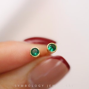 Puede incluir: Un par de pendientes de botón pequeños y redondos con piedras preciosas verdes vibrantes engastadas en oro. Los pendientes se muestran sobre un fondo suave y borroso, destacando el color y el diseño de la joyería.