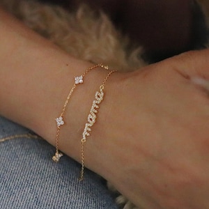 May include: Two gold bracelets are displayed on a wrist. One features a delicate chain with four-leaf clover charms. The other bracelet spells out "mama" in cursive with sparkling stones. The bracelets are set against a denim background.