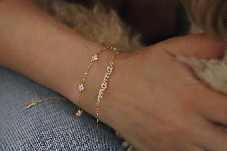 May include: Two gold bracelets are displayed on a wrist. One features a delicate chain with four-leaf clover charms. The other bracelet spells out "mama" in cursive with sparkling stones. The bracelets are set against a denim background.