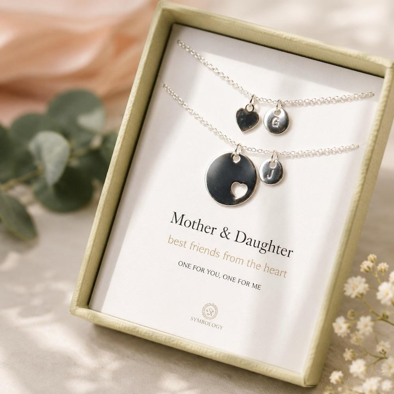 May include: Two silver-toned necklaces in a gift box. One features a small heart charm and a circular disc with the letter G. The other has a larger circular disc with a heart cutout and a smaller disc with the letter J. Text reads Mother & Daughter.