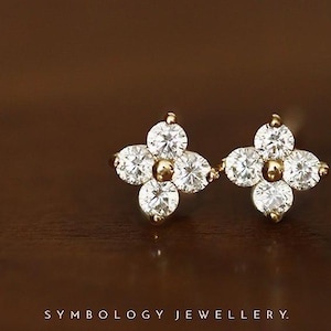 May include: A pair of gold stud earrings, each featuring four round clear stones arranged in a flower-like cluster with a central gold bead. The text SYMBOLOGY JEWELLERY is printed at the bottom of the frame in a clean, sans-serif font.