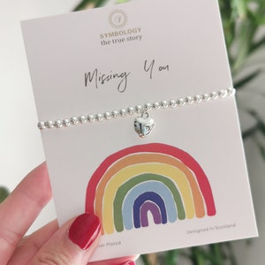 Puede incluir: Un brazalete plateado con un dije en forma de corazón y las palabras "Missing You" en una tarjeta blanca con un diseño de arcoíris. La tarjeta también dice "Designed in Scotland" y "Silver Plated".