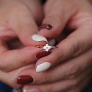 以下が含まれることがあります： 小さな透明な宝石がセットされた、四つ葉のクローバーのデザインのゴールドリング。 マニキュアされた白とバーガンディの色合いの爪を持つ2つの手でリングが保持されています。