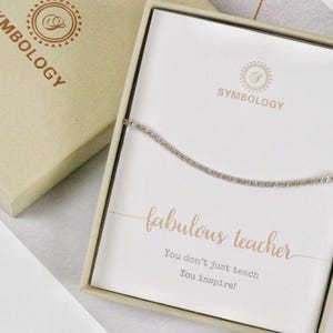 Könnte beinhalten: Eine weiße Geschenkbox mit einer silbernen Kettenkette darin. Die Box hat ein goldenes Logo mit der Aufschrift "Symbology" und eine Karte mit dem Text "fabulous teacher You don't just teach You inspire!"