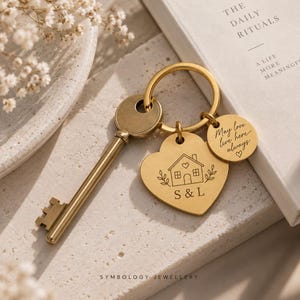 May include: A gold-toned metal key ring featuring a heart-shaped charm engraved with a house illustration, the letters S & L, and a smaller circular charm inscribed with the text May love live here always with a small heart symbol included.