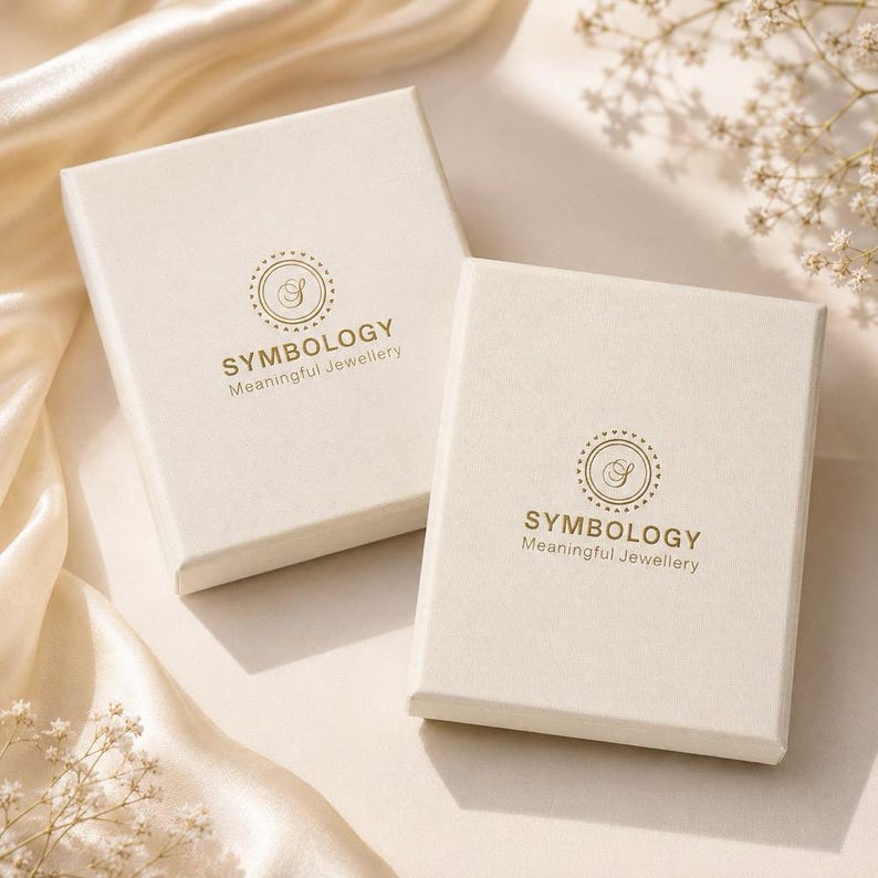 May include: Two rectangular off-white jewelry boxes featuring a gold circular logo with the letter S in the center, surrounded by the text SYMBOLOGY Meaningful Jewellery. The boxes are positioned on a smooth, light-colored fabric surface.