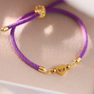 May include: A purple cord bracelet with gold-colored accents. The bracelet features a heart-shaped charm with a wing design. The adjustable bracelet is presented in a light-colored box.