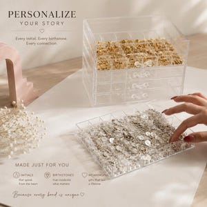 May include: A clear acrylic multi-drawer jewelry organizer containing small gold and silver circular charms. The text on the image reads: PERSONALIZE YOUR STORY, Every initial. Every birthstone. Every connection. MADE JUST FOR YOU.