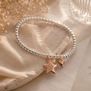 Op de afbeelding: Een elastische armband van kleine zilverkleurige kraaltjes met twee bungelende bedels: een roségoudkleurige ster met het ingegraveerde getal 18 en een klein roségoudkleurig hartje. De armband is gemaakt van uniforme metalen kralen op elastiek.