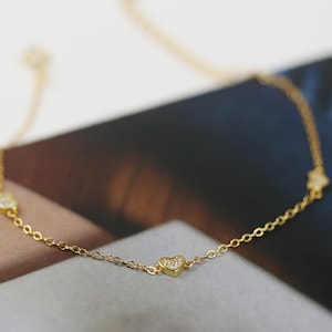 Może przedstawiać: Delikatny złoty łańcuszek z trzema małymi serduszkami, z których każde ozdobione jest błyszczącymi, przezroczystymi cyrkoniami.