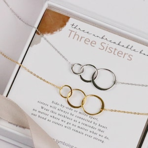Könnte beinhalten: Zwei silberne und goldene Halsketten mit ineinandergreifenden Kreisanhängern. Die Halsketten werden in einer weißen Schachtel mit einer Karte präsentiert, auf der "three unbreakable bonds. Three Sisters" steht und eine Botschaft über Schwestern, die durch das Herz verbunden sind, egal was passiert.