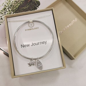 May include: A silver bangle bracelet with a charm that says "New Journey" in a box with the words "Symbology" and "Life is beautiful".