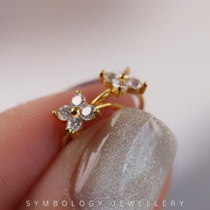 May include: A gold-colored double-flower earring with clear, sparkling gemstones. The earring is held by a person's hand, with the brand name "SYMBOLOLOGY JEWELLERY" visible at the bottom. The earring's design features two four-petal flower shapes.