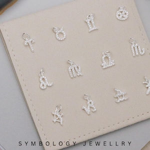 Puede incluir: Una colección de colgantes con dijes de signos del zodiaco plateados, cada uno incrustado con pequeñas piedras brillantes. Los dijes se exhiben sobre una superficie beige rectangular. La marca "SYMBOLOLOGY JEWELLRY" está impresa en negro en la parte inferior.