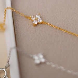 May include: Close-up of a gold necklace with a delicate chain and four-leaf clover shaped pendants. The clover pendants are adorned with small, sparkling white gemstones. A silver necklace with a similar design is also visible.