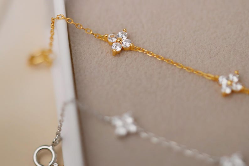 May include: Close-up of a gold necklace with a delicate chain and four-leaf clover shaped pendants. The clover pendants are adorned with small, sparkling white gemstones. A silver necklace with a similar design is also visible.