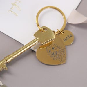 Può includere: Un set portachiavi color oro con una chiave, un ciondolo a forma di cuore con un'illustrazione di cane e un piccolo tag rotondo con il nome ".atte". Il portachiavi è su una superficie grigio chiaro.