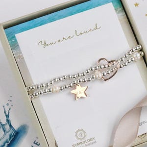 Op de afbeelding: Een zilveren en roségouden armband met een hartbedel en een sterbedel met de initialen "R & S" erop gegraveerd. De armband zit in een doosje met een kaartje waarop "You are loved" staat.