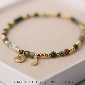 May include: A beaded bracelet with green and gold beads, accented with gold-coloured spacers. The bracelet features two gold charms, one shaped like the letter "D" and the other "J", both encrusted with small clear stones. The bracelet is presented in a white box.
