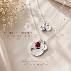 May include: Two silver necklaces featuring circular pendants and charms. One pendant has a heart cutout and a red stone, the other a purple stone. Both have small round charms engraved with the letter J. Text reads MOTHER & DAUGHTER CONNECTED BY HEART.