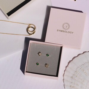 May include: A gold necklace with interlocking circles and a pair of stud earrings in a pink box. The earrings include a pair of star-shaped studs and a pair of green gemstone studs. The box is labeled "SYMBOLOLOGY".